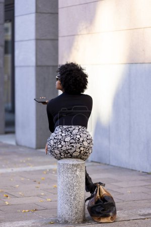 Téléchargez les photos : Femme afro très sexy assis sur un pieu en ville est au téléphone levant les yeux. Sur le sol est son sac à main. Scène urbaine. - en image libre de droit
