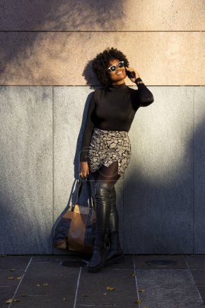 Téléchargez les photos : Sexy femme afro courbée est appuyé contre un mur et est au téléphone souriant et levant les yeux. Elle tient son sac. Elle est éclairée par la lumière directe du soleil. - en image libre de droit