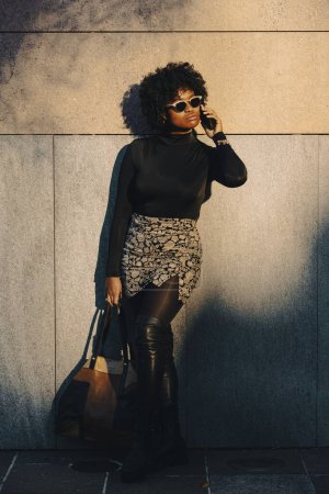 Téléchargez les photos : Sexy femme afro courbée est appuyé contre un mur et est sur son téléphone regardant vers le bas. Elle tient son sac. Elle est éclairée par la lumière directe du soleil. - en image libre de droit