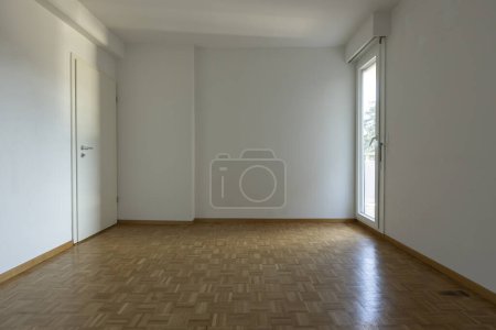 Téléchargez les photos : Vue de face d'une chambre vide avec parquet et murs blancs. Sur la gauche se trouve une fenêtre laissant entrer beaucoup de lumière naturelle, sur la droite se trouve une porte fermée. Personne à l'intérieur. - en image libre de droit