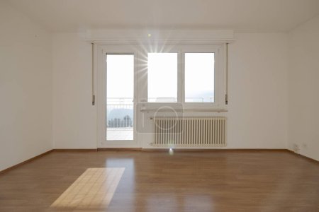 Téléchargez les photos : Chambre vide avec parquet, sous la fenêtre il y a un grand radiateur. De la porte, vous pouvez sortir sur le petit balcon. Personne à l'intérieur - en image libre de droit