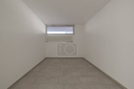 Téléchargez les photos : Vue de face d'une petite chambre vide avec des murs blancs une petite fenêtre et le radiateur suspendu au mur. Personne à l'intérieur - en image libre de droit