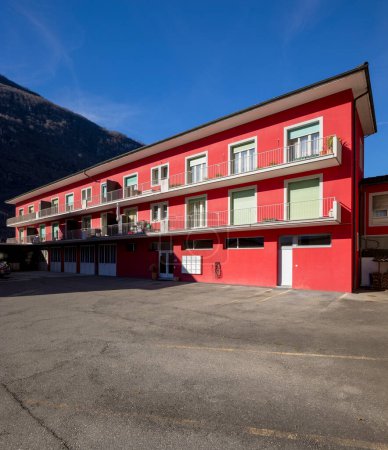 Photo for Red railing house with parking area below. Some windows open, others closed by green shutters. Blue sky and mountains behind. Nobody inside - Royalty Free Image