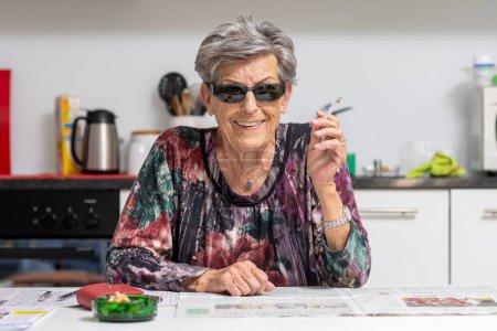 Foto de Una anciana con arrugas está sentada en la cocina usando gafas de sol mientras fuma alegremente un cigarrillo.. - Imagen libre de derechos