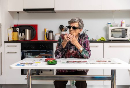 Elderly lady with dark glasses, lights a cigarette in the kitchen in front of her newspaper and ashtray.