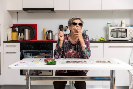 Photo for Old lady full of wrinkles smokes comfortably at home in her kitchen wearing black sunglasses. - Royalty Free Image