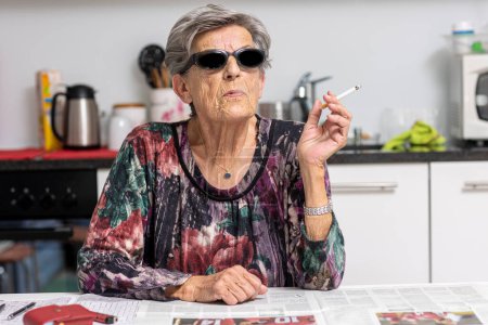 Téléchargez les photos : Portrait d'une vieille fumeuse assise dans sa cuisine blanche moderne avec des lunettes de soleil sombres. Une femme d'une autre époque. - en image libre de droit