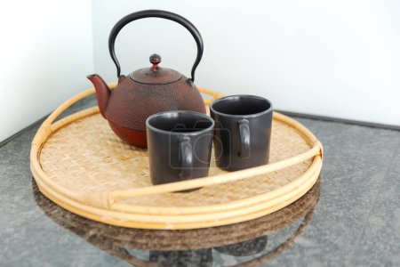 Téléchargez les photos : Détail d'une théière chinoise rouge et de deux tasses noires reposant sur un plateau en osier, dans une cuisine lumineuse. Personne à l'intérieur - en image libre de droit