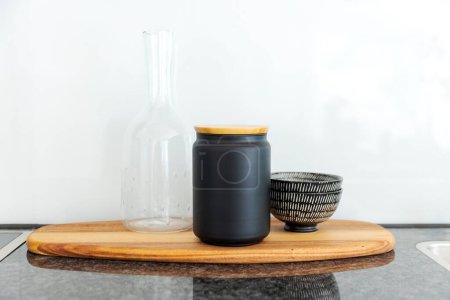 Photo for Front view of an elegant wooden tray with a black vase with wooden lid, a glass pitcher and a bowl resting on it. Elegant and minimalist - Royalty Free Image