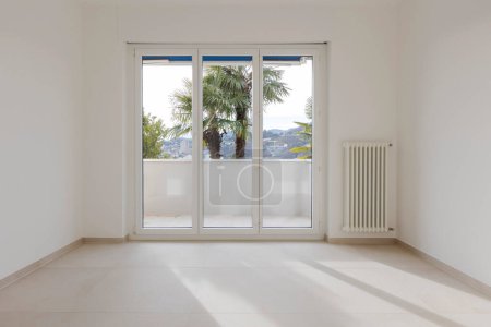 Foto de Habitación vacía con una gran ventana en el fondo que conduce al balcón. Un radiador o calentador se puede ver a la derecha en la pared blanca. - Imagen libre de derechos
