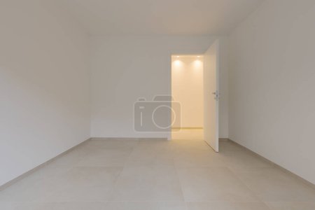 Photo for Inside an empty room and to the right a door leading to the corridor with a light on. All the walls and ceiling are white. - Royalty Free Image