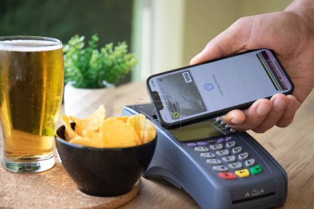 Photo for Alanya, Turkey - October 4, 2022: Man hand holding iPhone 14 with app Raiffeisenbank on the screen and pay pass online POS terminal buy the Beer in the cafe. - Royalty Free Image