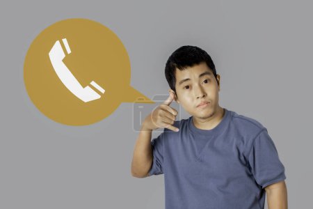 Téléchargez les photos : Portrait of young single man wearing t-shirt making call me gesture with icon telephone, sign with hand shaped like phone isolated on gray background. Positive human emotions, face expressions. - en image libre de droit