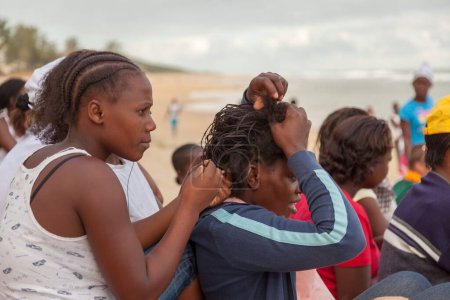 Téléchargez les photos : Praia de Xai Xai, Mozambique, avril 2014 : Jeunes adolescentes africaines tressant des cheveux en rangs de maïs alors qu'elles étaient assises sur le sable à Praia de Xai Xai à l'ombre, prêtes à regarder un match amical de soccor sur la plage après le culte religieux pentecôtiste du dimanche. Allez-y 2 M - en image libre de droit