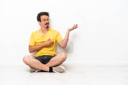 Téléchargez les photos : Jeune homme caucasien assis sur le sol isolé sur fond blanc étendant les mains sur le côté pour inviter à venir - en image libre de droit