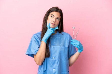 Téléchargez les photos : Dentiste femme tenant des outils isolés sur fond rose ayant des doutes tout en levant les yeux - en image libre de droit