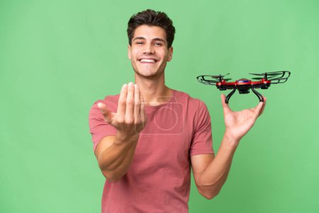 Junger kaukasischer Mann, der eine Drohne über isoliertem Hintergrund hält und einlädt, mit der Hand zu kommen. Glücklich, dass du gekommen bist