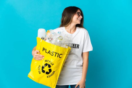 Photo for Woman holding a bag full of bottles to recycle isolated on blue background laughing in lateral position - Royalty Free Image