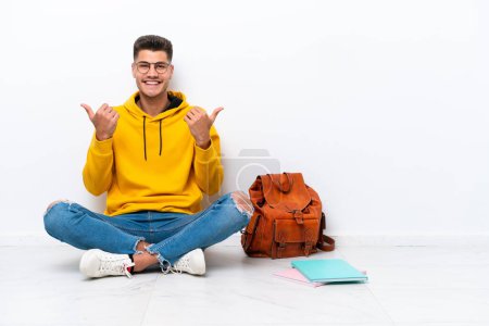 Photo for Young student caucasian man sitting one the floor isolated on white background with thumbs up gesture and smiling - Royalty Free Image