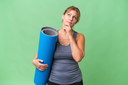 Middle-aged sport woman going to yoga classes while holding a mat over isolated background and looking up