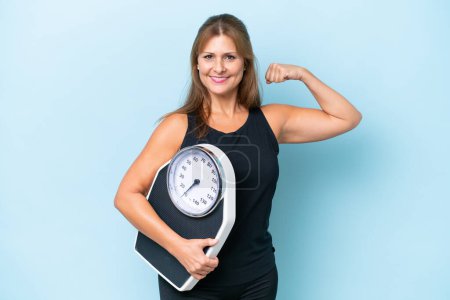 Photo for Middle-aged caucasian woman isolated on blue background holding a weighing machine and doing strong gesture - Royalty Free Image