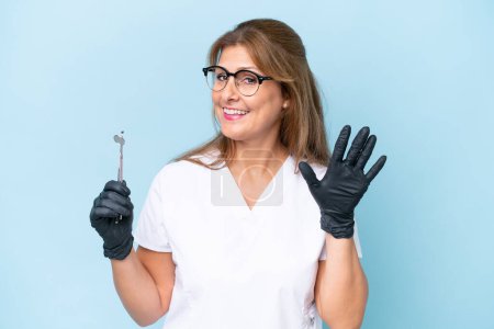 Foto de Dentista de mediana edad sosteniendo herramientas sobre fondo azul aislado saludando con la mano con expresión feliz - Imagen libre de derechos