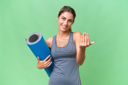 Pretty Young Uruguayan sport woman going to yoga classes while holding a mat over isolated background inviting to come with hand. Happy that you came