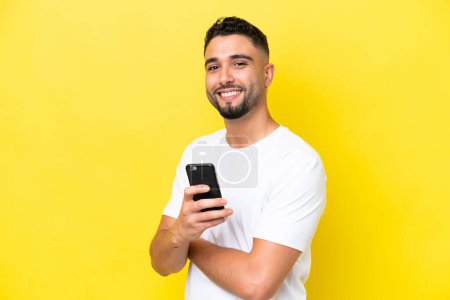 Jeune homme arabe beau isolé sur fond jaune tenant un téléphone portable et les bras croisés