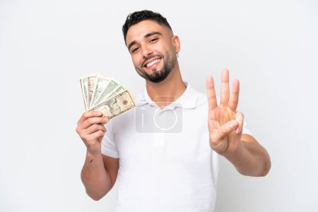Young Arab man taking a lot of money isolated on white background happy and counting three with fingers