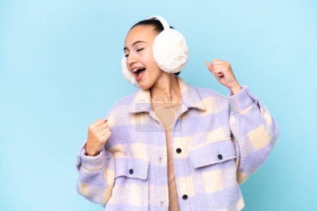 Young Arab woman wearing winter muffs isolated on blue background celebrating a victory