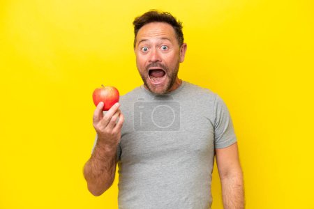 Foto de Hombre caucásico de mediana edad con una manzana aislada sobre fondo amarillo con expresión facial sorpresa y conmocionada - Imagen libre de derechos