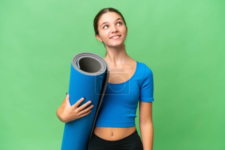 Teenager caucasian girl going to yoga classes while holding a mat over isolated background looking up while smiling