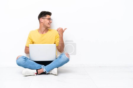 Foto de Young man sitting on the floor isolated on white background pointing to the side to present a product - Imagen libre de derechos