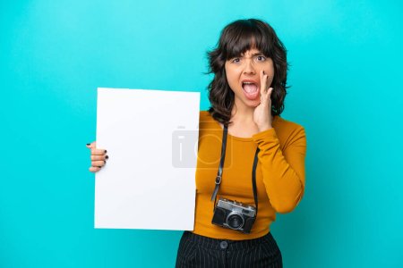 Junge Fotografin lateinische Frau isoliert auf blauem Hintergrund hält ein leeres Plakat und schreit