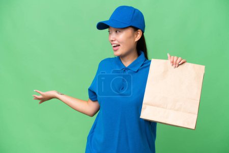 Foto de Joven mujer asiática tomando una bolsa de comida para llevar sobre un fondo aislado con expresión sorpresa mientras mira al lado - Imagen libre de derechos