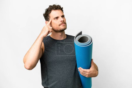 Young sport man going to yoga classes while holding a mat over isolated white background having doubts and thinking