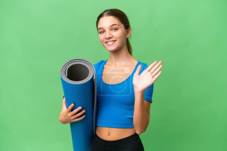 Teenager caucasian girl going to yoga classes while holding a mat over isolated background saluting with hand with happy expression