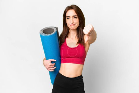 Téléchargez les photos : Young sport caucasian woman going to yoga classes while holding a mat over isolated white background showing thumb down with negative expression - en image libre de droit