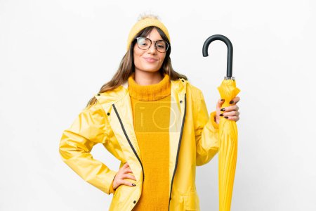 Téléchargez les photos : Young caucasian woman with rainproof coat and umbrella over isolated white background posing with arms at hip and smiling - en image libre de droit