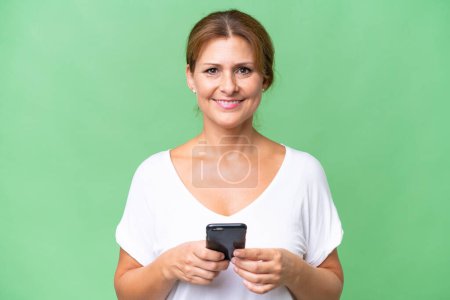 Photo for Middle-aged caucasian woman over isolated background looking at the camera and smiling while using the mobile - Royalty Free Image