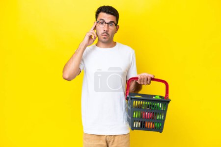 Photo for Young handsome man holding a shopping basket full of food over isolated yellow background thinking an idea - Royalty Free Image