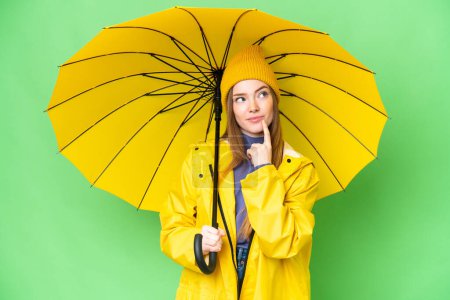 Téléchargez les photos : Jeune jolie femme avec manteau imperméable et parapluie sur fond chromatique isolé ayant des doutes tout en levant les yeux - en image libre de droit