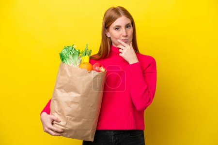Foto de Mujer pelirroja joven sosteniendo una bolsa de la compra de comestibles aislado en el pensamiento de fondo amarillo - Imagen libre de derechos