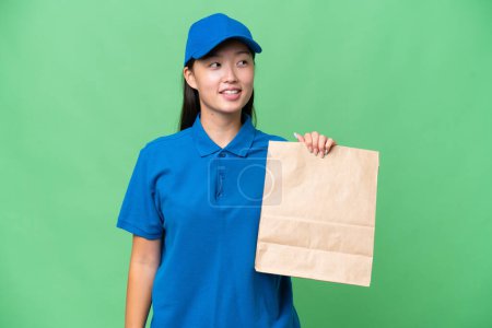 Foto de Young Asian woman taking a bag of takeaway food over isolated background looking side - Imagen libre de derechos