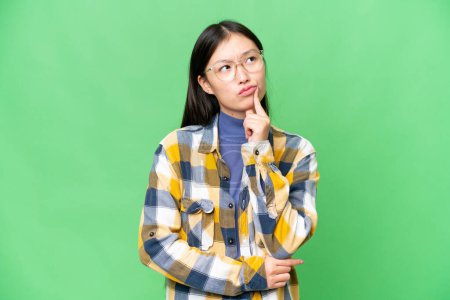 Foto de Young Asian woman over isolated chroma key background having doubts while looking up - Imagen libre de derechos