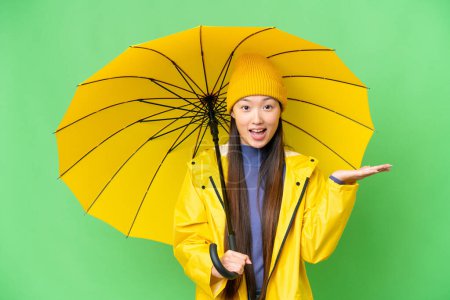Téléchargez les photos : Young Asian woman with rainproof coat and umbrella over isolated chroma key background with shocked facial expression - en image libre de droit
