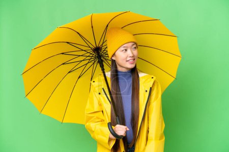 Téléchargez les photos : Young Asian woman with rainproof coat and umbrella over isolated chroma key background looking to the side and smiling - en image libre de droit