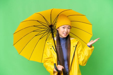 Téléchargez les photos : Young Asian woman with rainproof coat and umbrella over isolated chroma key background extending hands to the side for inviting to come - en image libre de droit