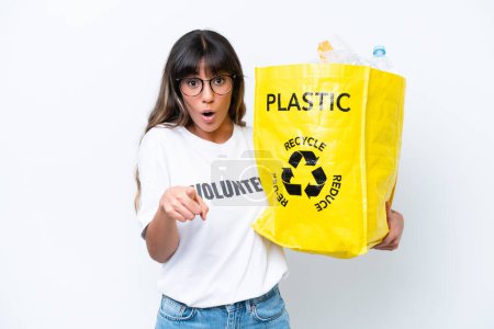 Foto de Joven mujer caucásica sosteniendo una bolsa llena de botellas de plástico para reciclar aislado sobre fondo blanco sorprendido y apuntando hacia delante - Imagen libre de derechos