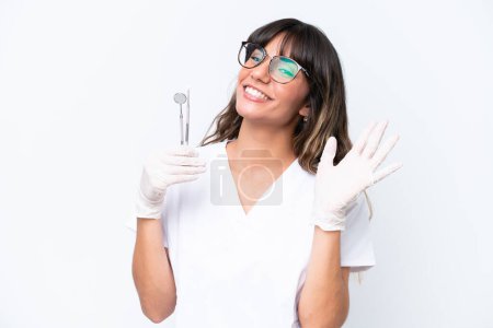 Foto de Dentista mujer caucásica sosteniendo herramientas aisladas sobre fondo blanco saludando con la mano con expresión feliz - Imagen libre de derechos